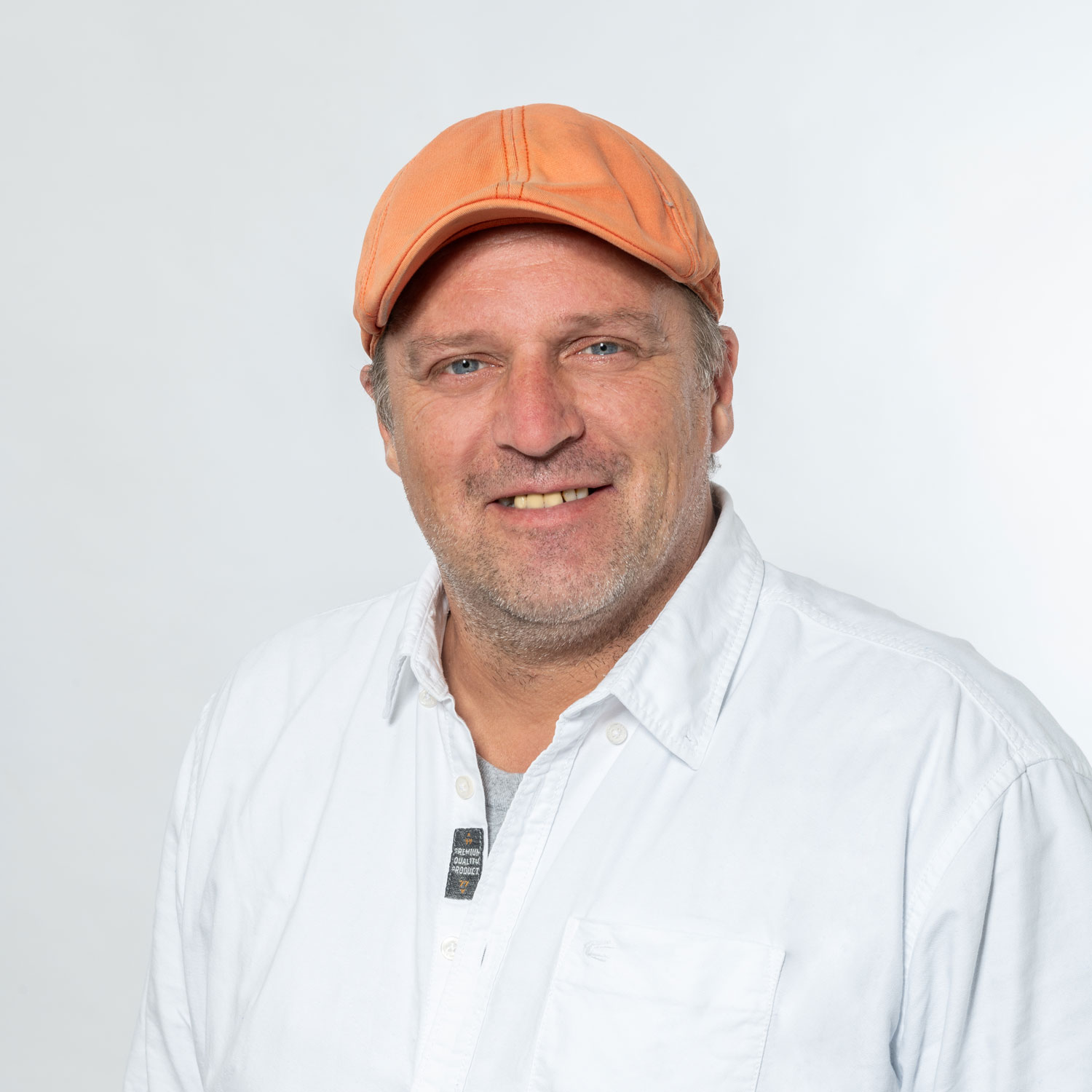 Mann mit orangefarbener Mütze und weißem Hemd, der in die Kamera lächelt, vor einem schlichten hellen Hintergrund.