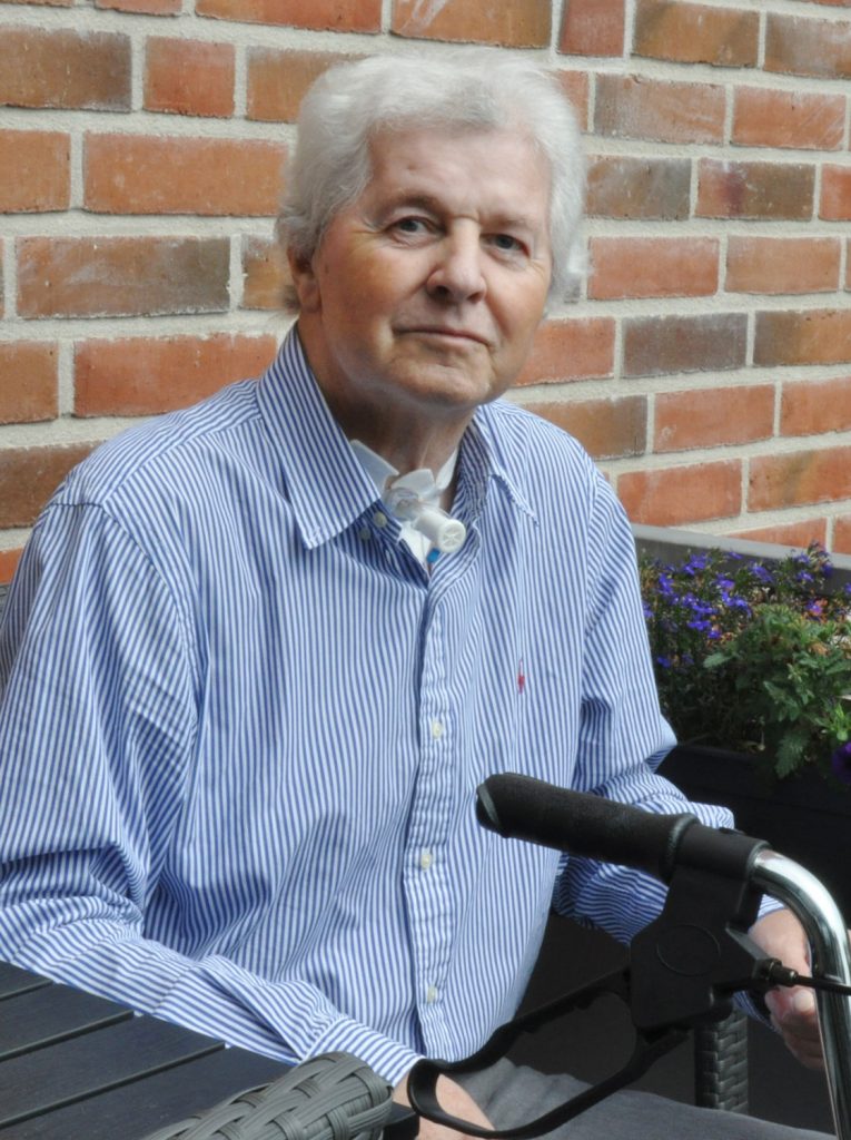 Ein älterer Mann mit weißem Haar und gestreiftem blauem Hemd sitzt an einem Tisch im Freien an einer Backsteinwand. An seinem Hals trägt er ein medizinisches Gerät und vor ihm liegt eine Gehhilfe.