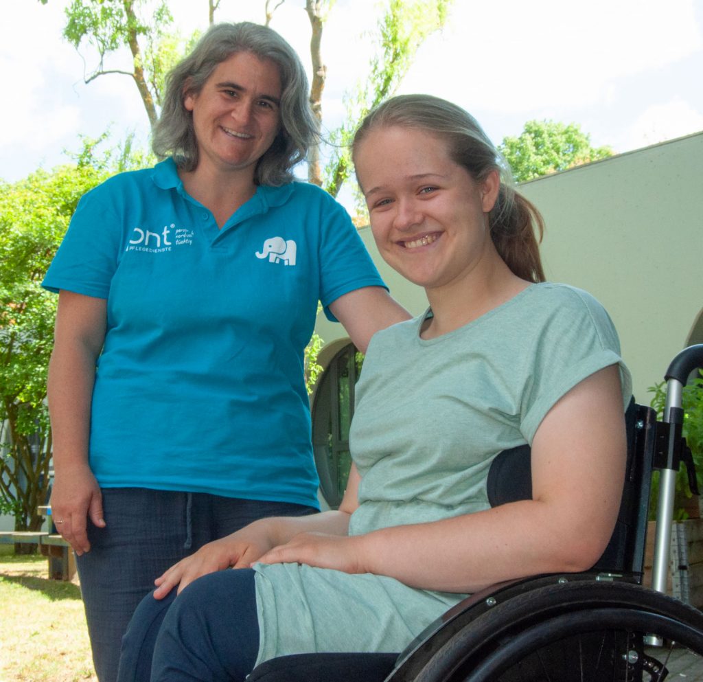 Zwei lächelnde Frauen im Freien; eine im Rollstuhl und eine andere daneben, beide in Freizeitkleidung. Mitarbeiterin vom Kinderpflegedienst Lübeck.