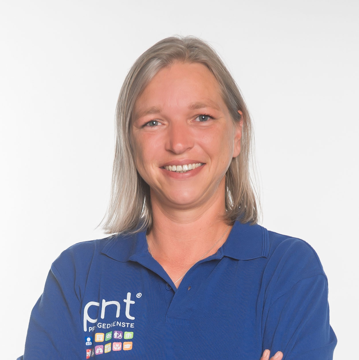 Eine Frau mit glattem blondem Haar, lächelnd, trägt ein blaues Poloshirt mit dem Aufdruck "pnt" und verschiedenen Symbolen und steht vor einem schlichten weißen Hintergrund.