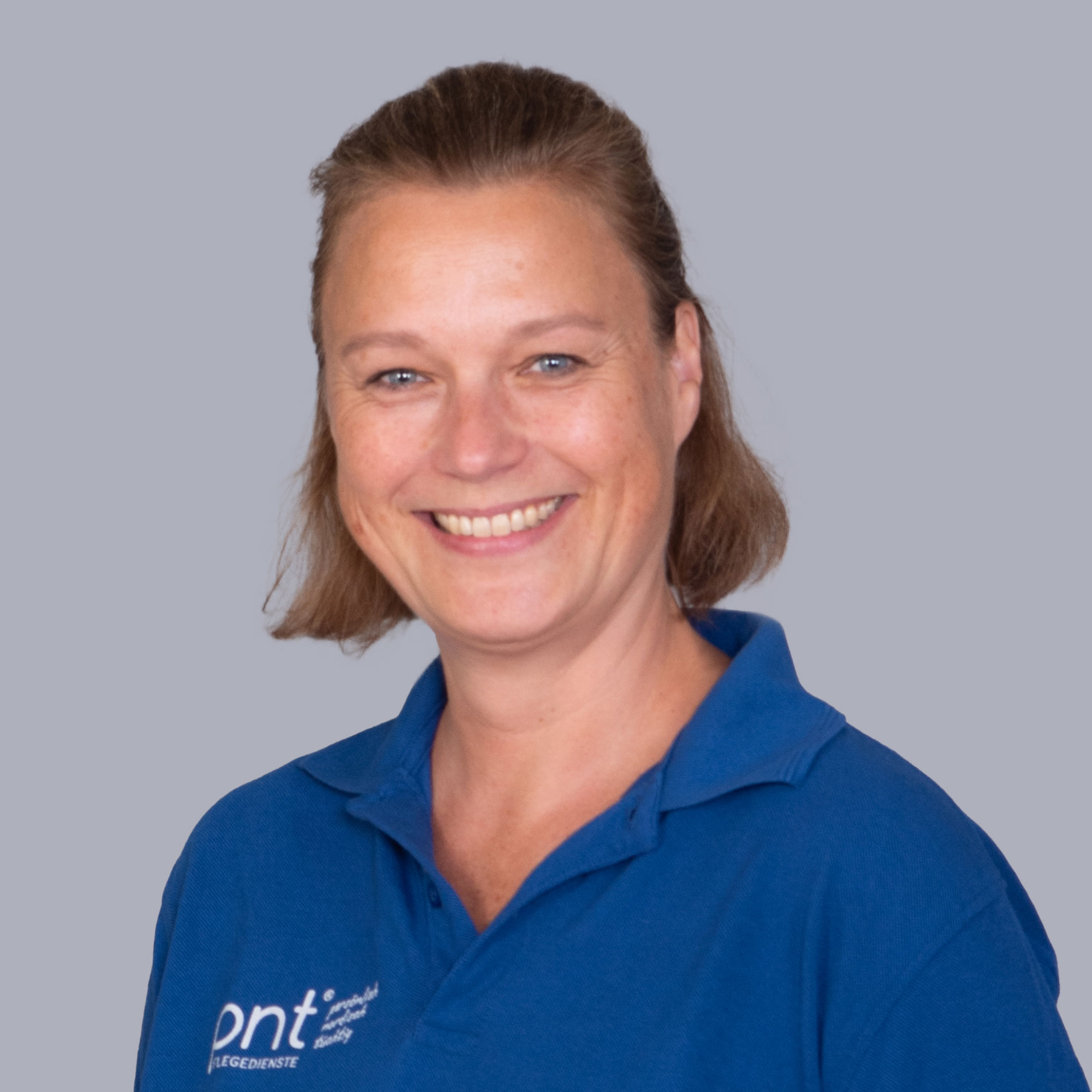 Eine Frau mit kurzen hellbraunen Haaren lächelt in die Kamera. Sie trägt ein blaues Hemd mit Kragen und weißem Logo und steht vor einem schlichten grauen Hintergrund.
