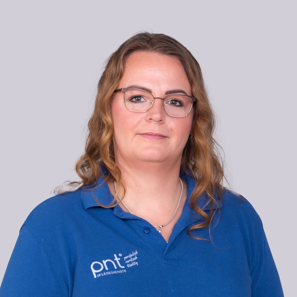 Eine Frau mit gewelltem braunem Haar und Brille, die ein blaues Kragenhemd mit einem "pnt"-Logo trägt, posiert vor einem schlichten hellgrauen Hintergrund.