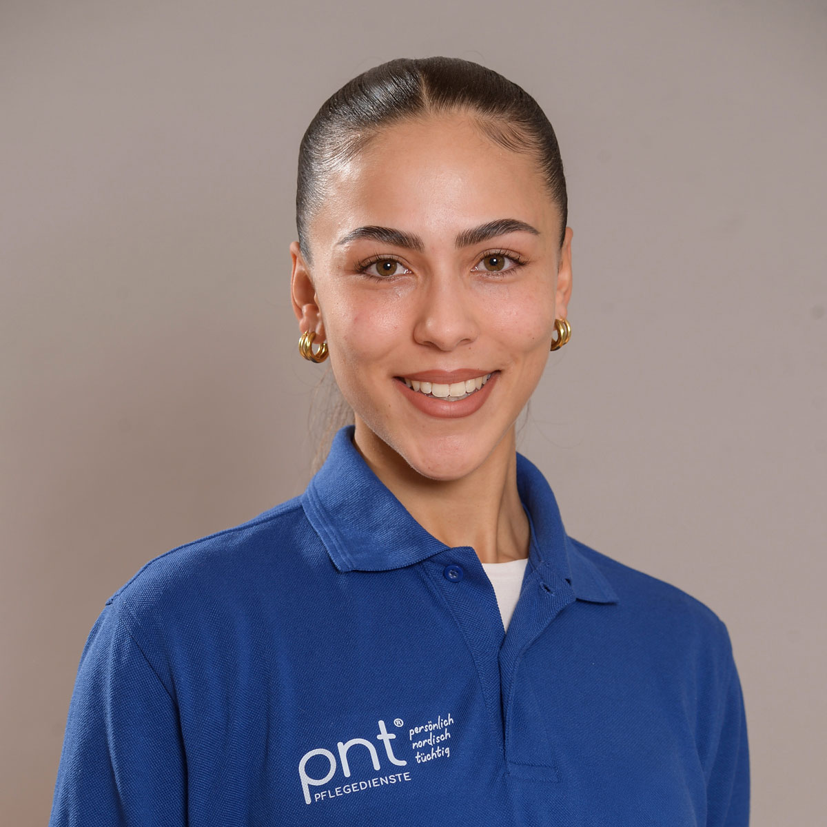 Frau in einem blauen "pnt Pflegedienste"-Poloshirt, lächelnd in die Kamera, mit zurückgestyltem Haar und goldenen Ohrringen, vor einem unifarbenen Hintergrund.