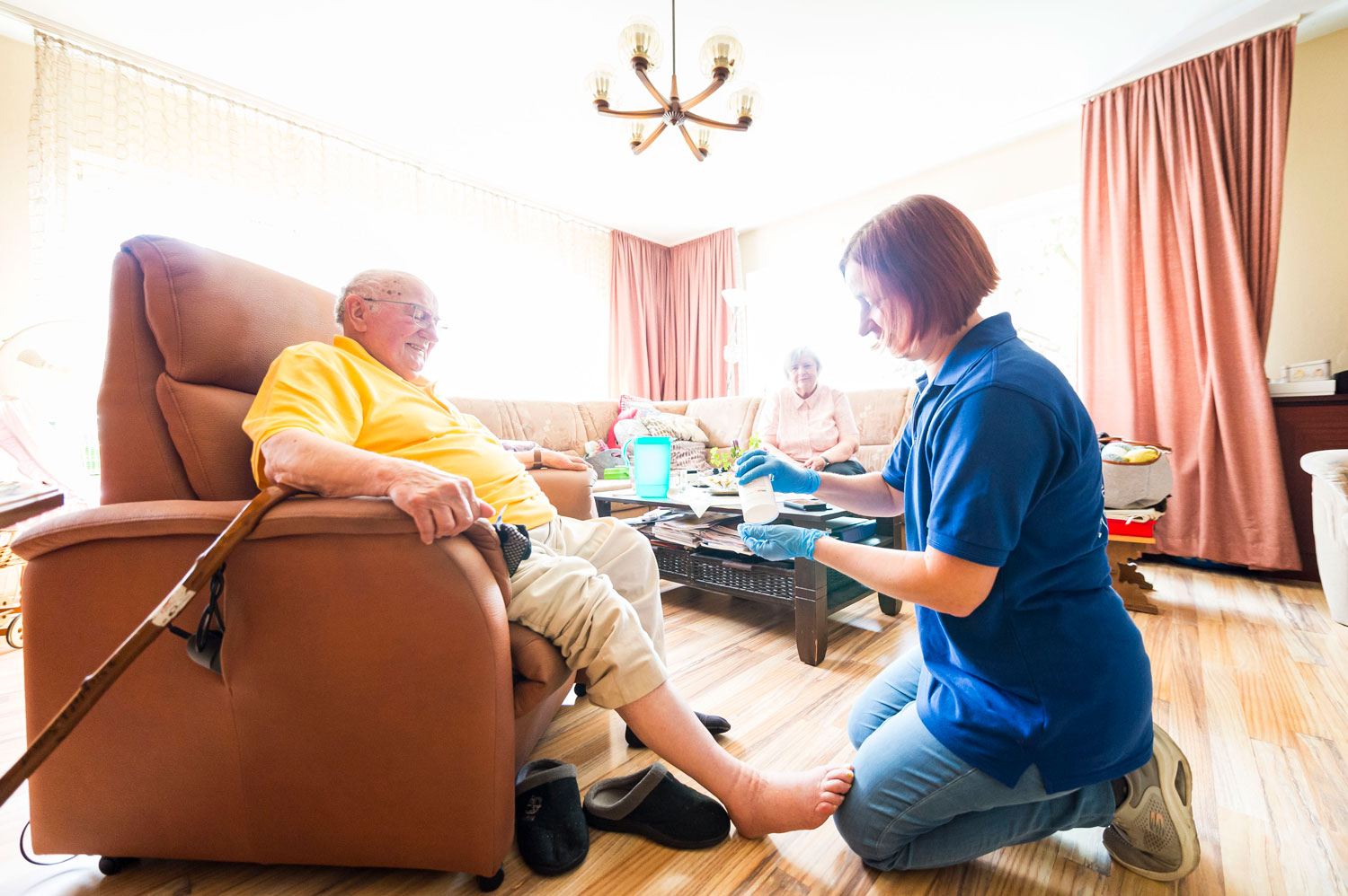 Eine Pflegekraft in blauer Uniform versorgt in einem hellen Wohnzimmer den Fuß eines älteren Mannes, während im Hintergrund eine ältere Frau auf dem Sofa sitzt.