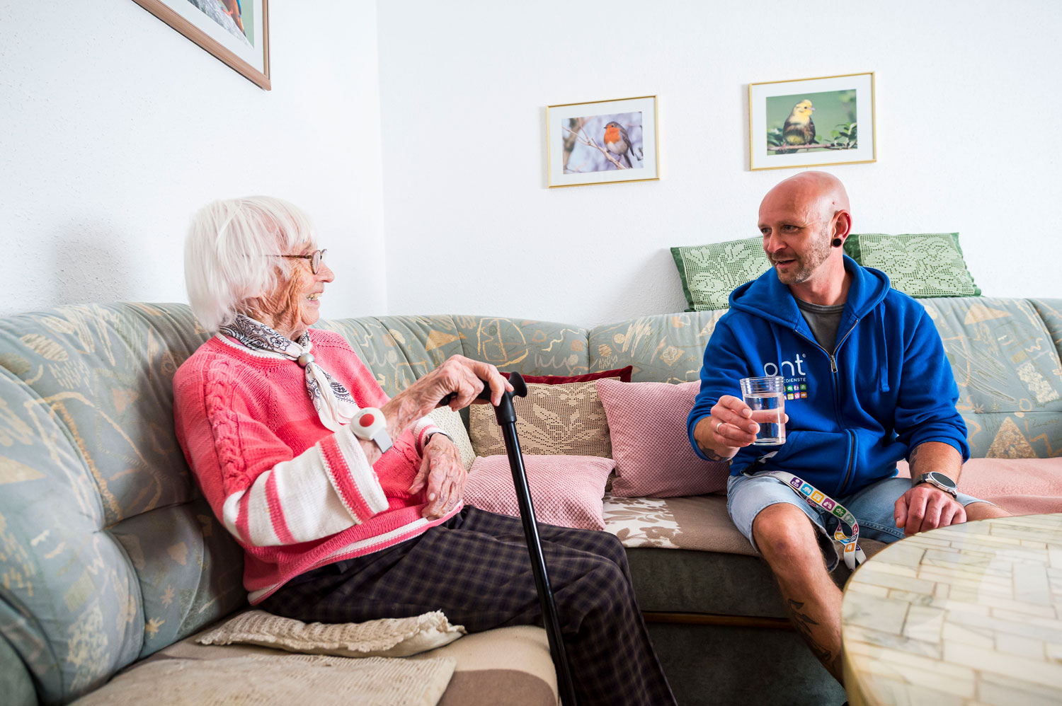 Eine ältere Frau mit einem Gehstock sitzt auf einem Sofa und unterhält sich mit einem Mann in einem blauen Kapuzenpulli, der ein Glas Wasser in der Hand hält, in einem hellen Wohnzimmer.