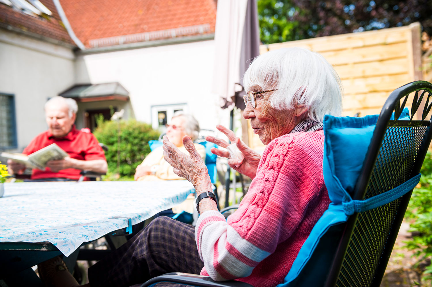 Eine ältere Frau sitzt an einem Tisch im Freien und unterhält sich, während ein anderer älterer Mann in der Nähe sitzt und Zeitung liest. Die Szene spielt in einem sonnigen Garten oder auf einer Terrasse.