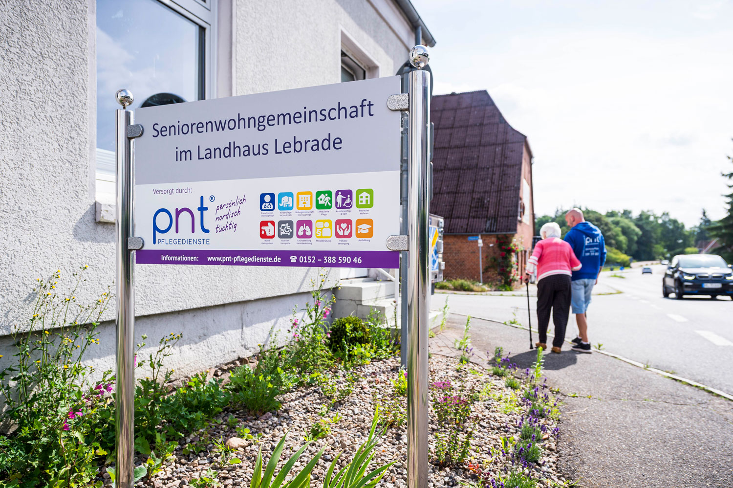 Am Gehweg steht ein Schild mit der Aufschrift „Seniorenwohngemeinschaft im Landhaus Lebrade“; im Hintergrund sind zwei ältere Menschen zu sehen, die gemeinsam in der Nähe einer Wohnstraße spazieren gehen.