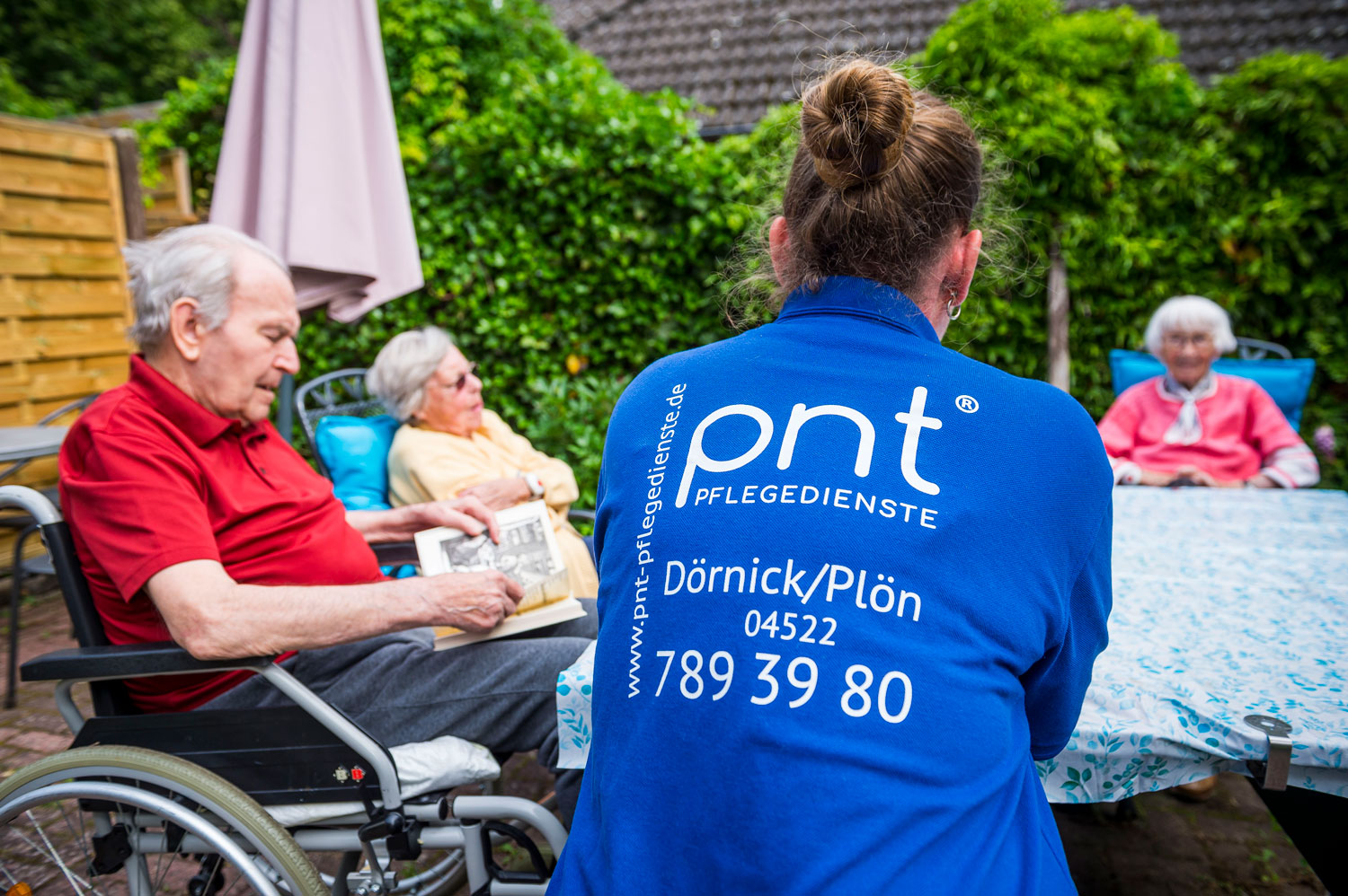 Eine Pflegekraft in einer blauen Jacke sitzt mit drei älteren Menschen, von denen zwei im Rollstuhl sitzen, an einem Tisch im Freien in einer Gartenanlage.