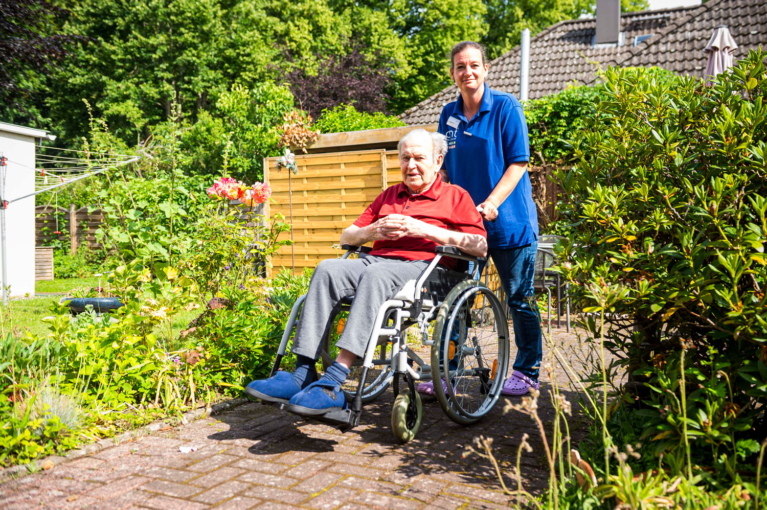 An einem sonnigen Tag wird ein älterer Mann im Rollstuhl von einer Pflegekraft auf einem Gartenweg geschoben.