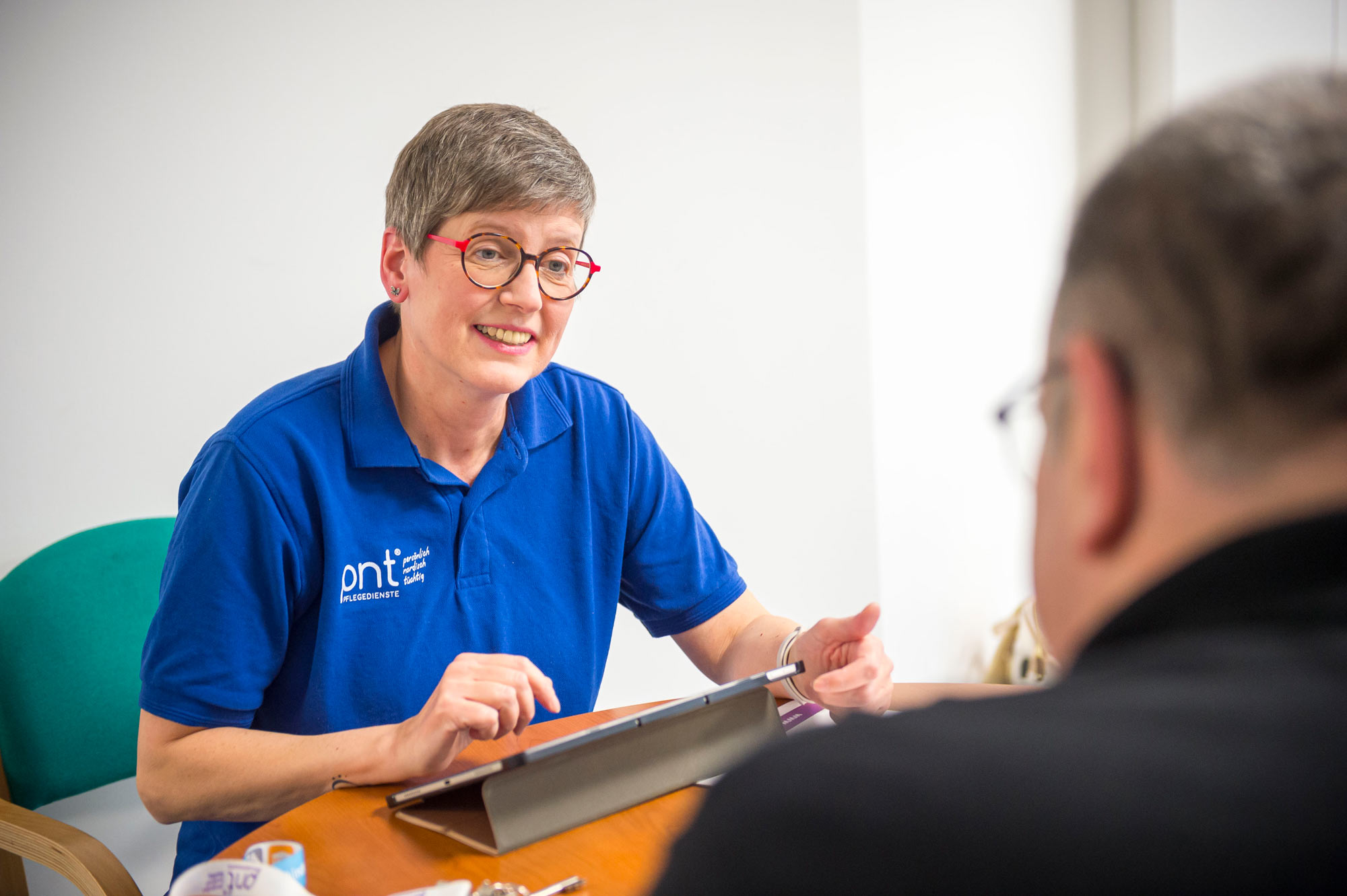 Eine Frau in einem blauen Poloshirt führt eine Pflegeberatung durch: Sie sitzt an einem Tisch, unterhält sich einfühlsam mit einem Mann gegenüber und macht sich Notizen auf einem Tablet.