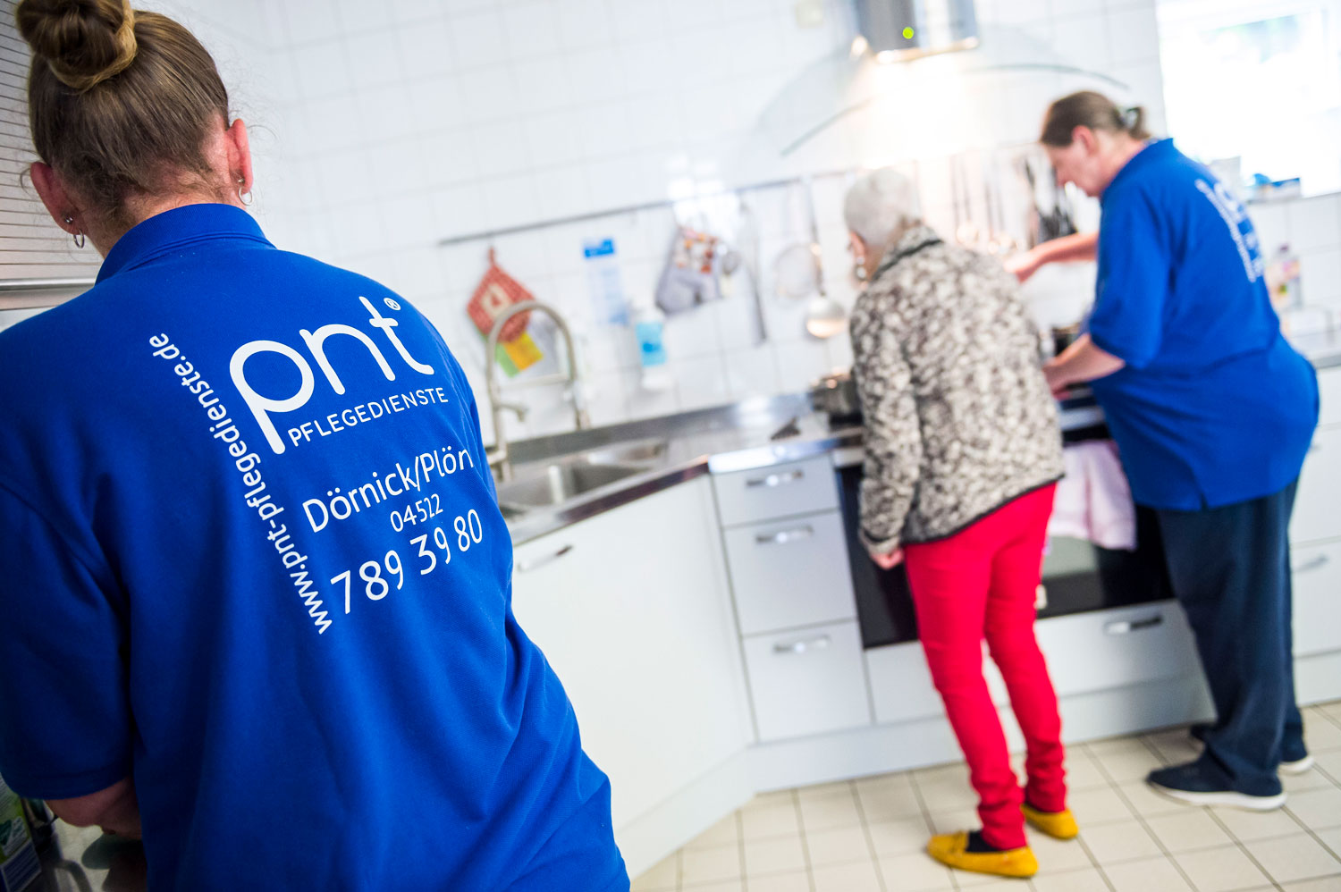 Zwei Pflegekräfte in blauen Uniformen helfen einer älteren Frau in der Küche; eine Pflegekraft putzt, während die andere beim Kochen hilft.