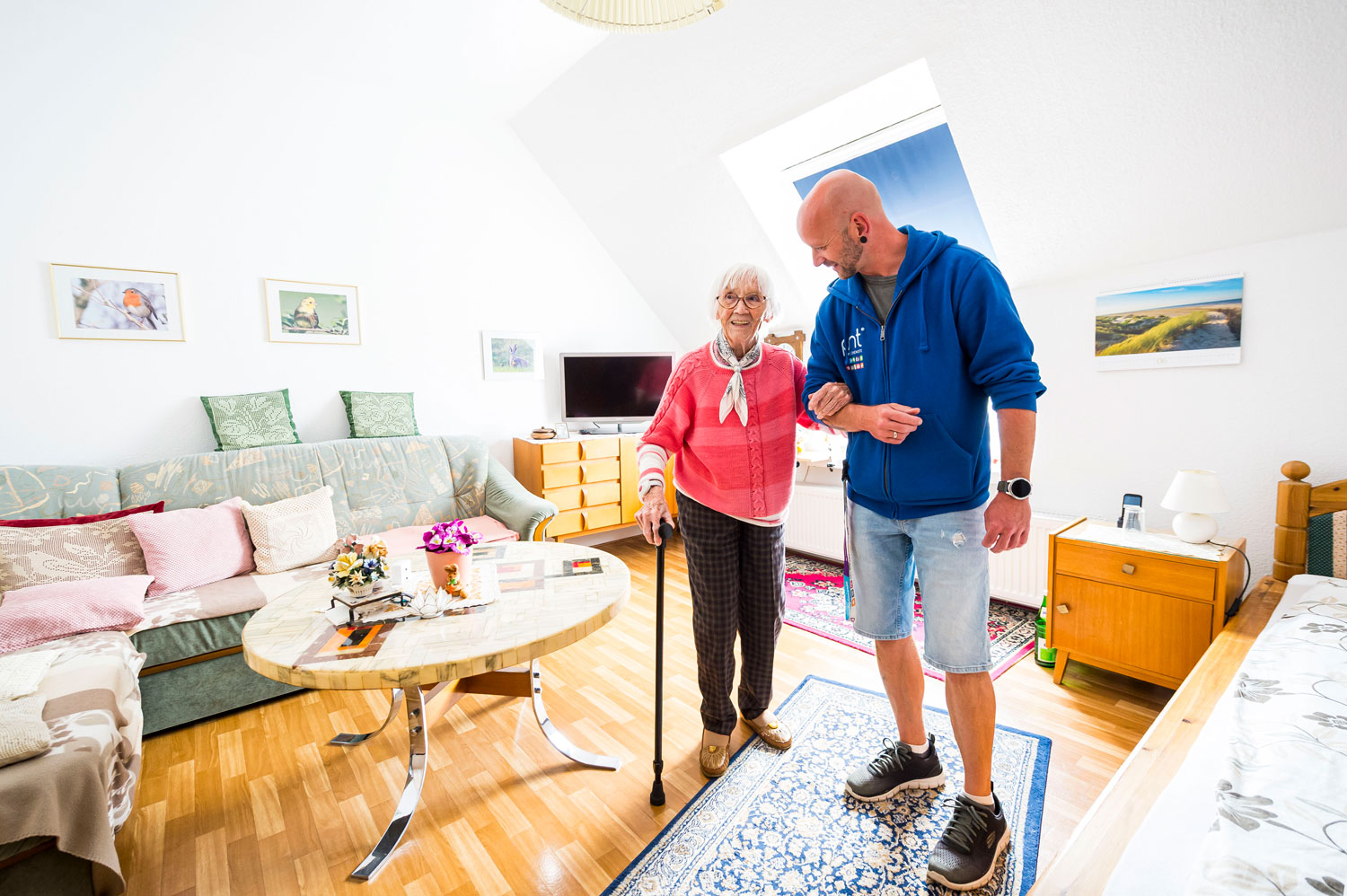 Eine ältere Frau mit einem Gehstock wird von einem Mann in einem blauen Freizeit-Hoodie in einem hellen, aufgeräumten Wohnzimmer mit Holzböden und gerahmten Fotos an den Wänden unterstützt.