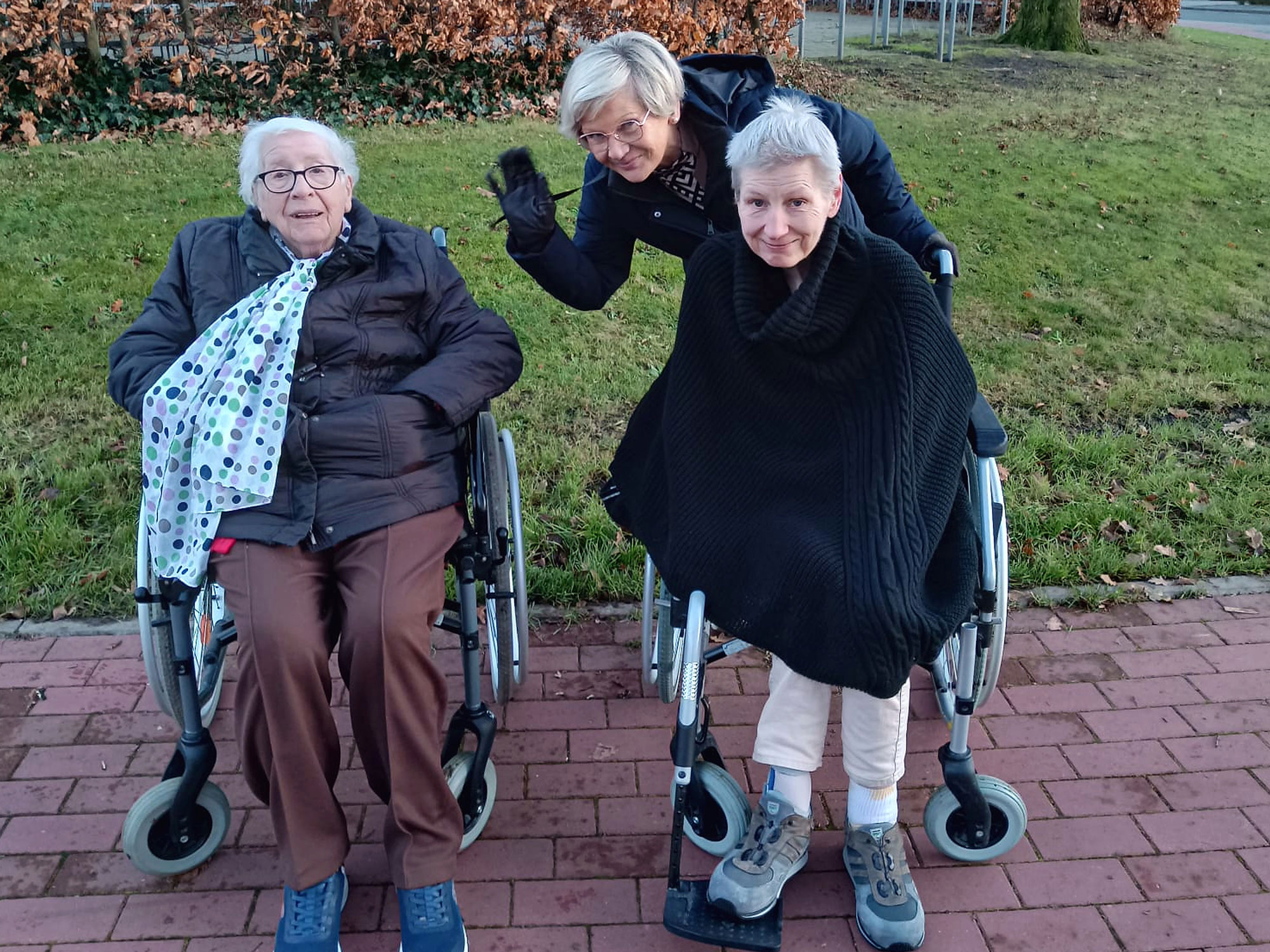 Zwei ältere Frauen sitzen in Rollstühlen auf einem gepflasterten Weg, hinter ihnen steht eine Frau, die winkt und lächelt. Im Hintergrund sind grünes Gras und Herbstblätter zu sehen.