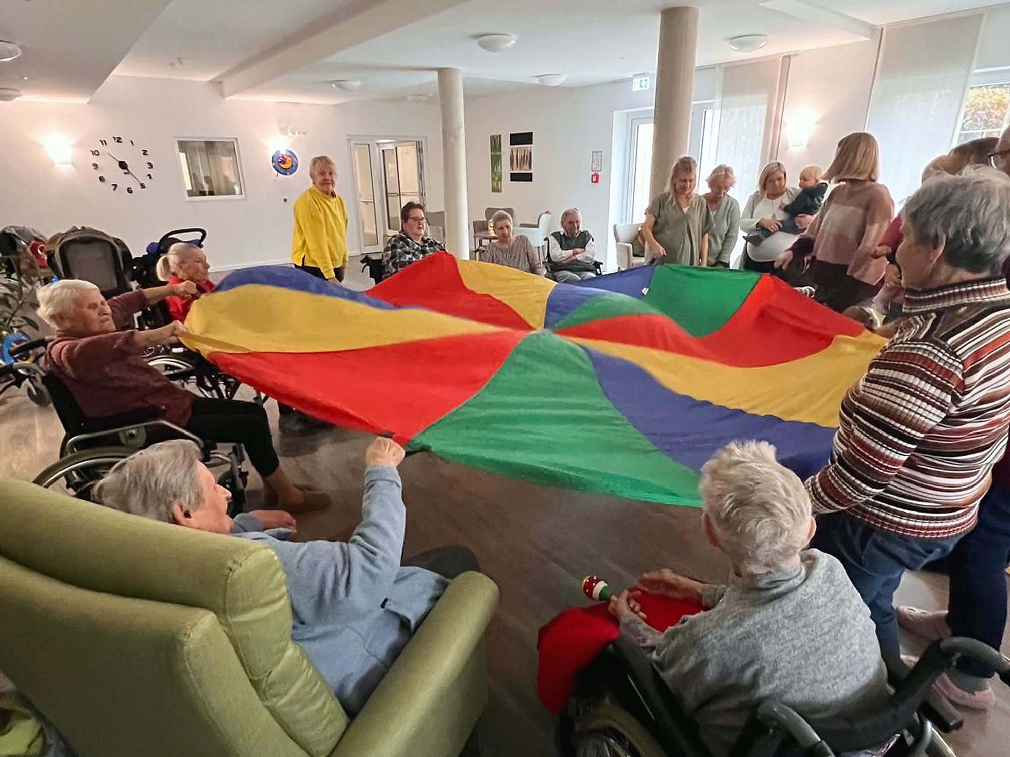 Eine Gruppe älterer Menschen und Pflegekräfte hält in einem Raum einen bunten Fallschirm hoch und nimmt an einer Gruppenaktivität in einer Pflegeeinrichtung teil.