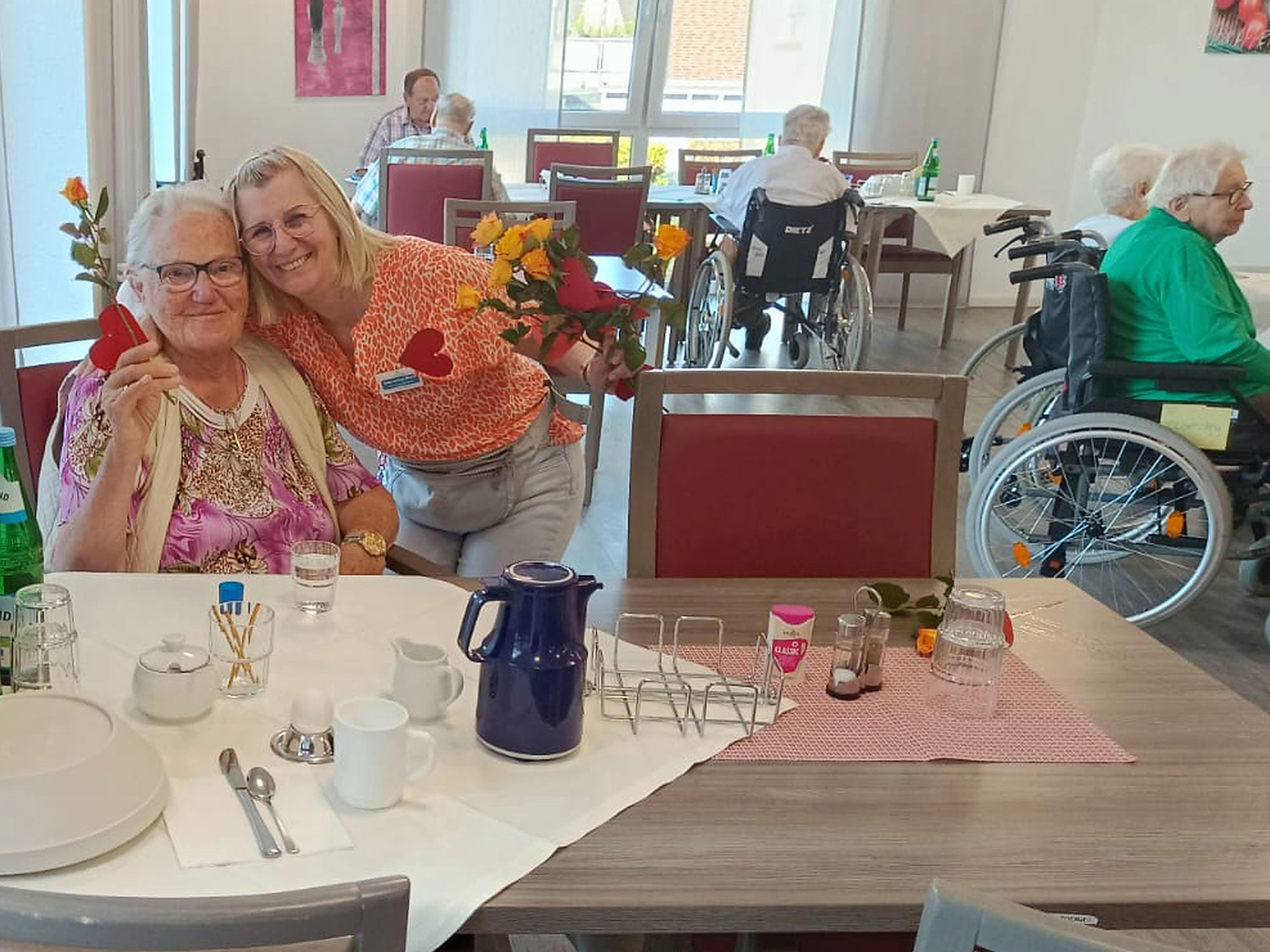 Zwei Frauen lächeln und posieren mit Blumen an einem Esstisch in einer Seniorenpflegeeinrichtung, während andere Bewohner an benachbarten Tischen und in Rollstühlen sitzen.