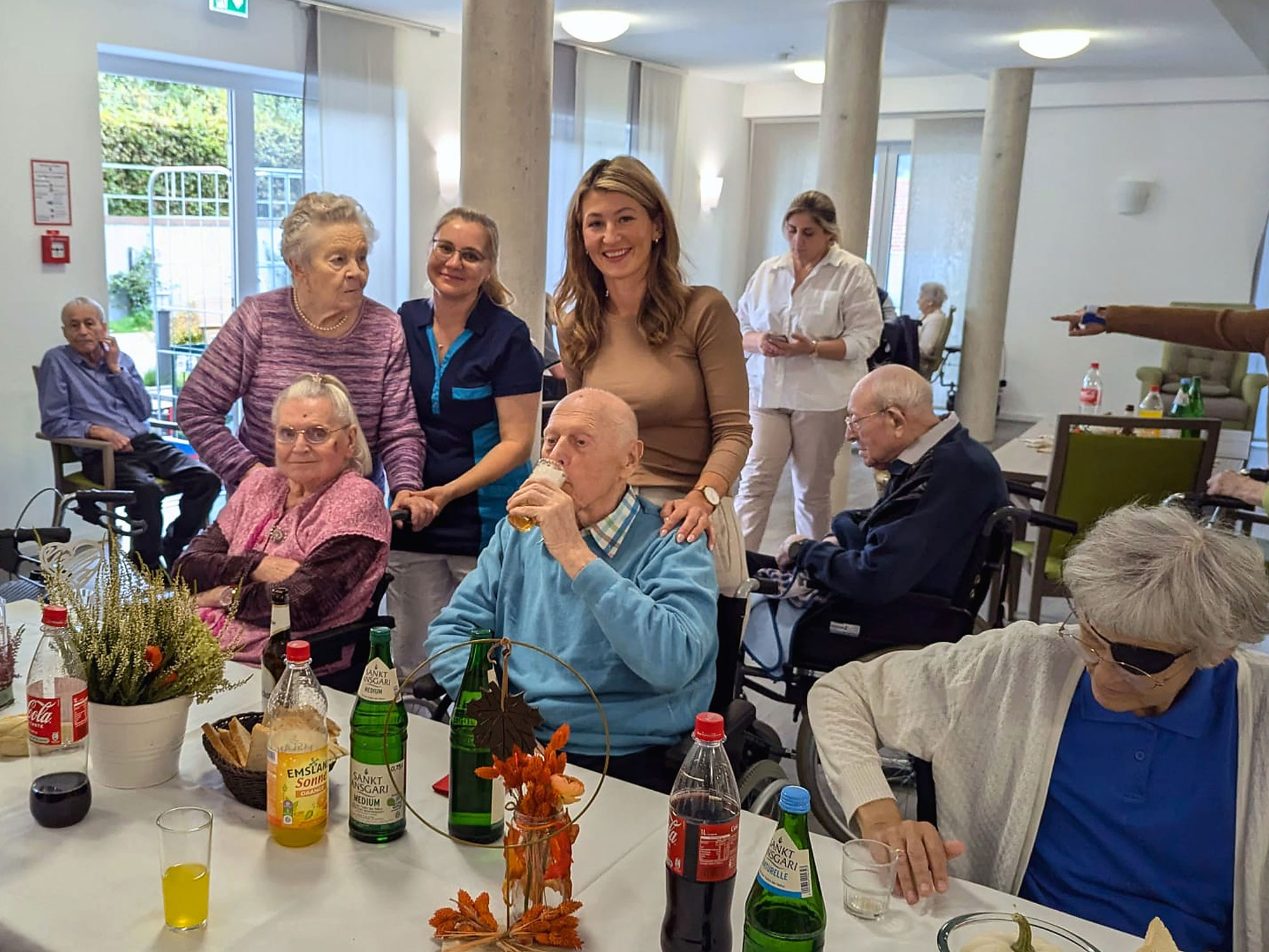 In einem hell erleuchteten Gemeinschaftsraum versammelt sich eine Gruppe älterer Menschen und Pflegekräfte um einen Tisch, auf dem Getränke und Dekorationen stehen.