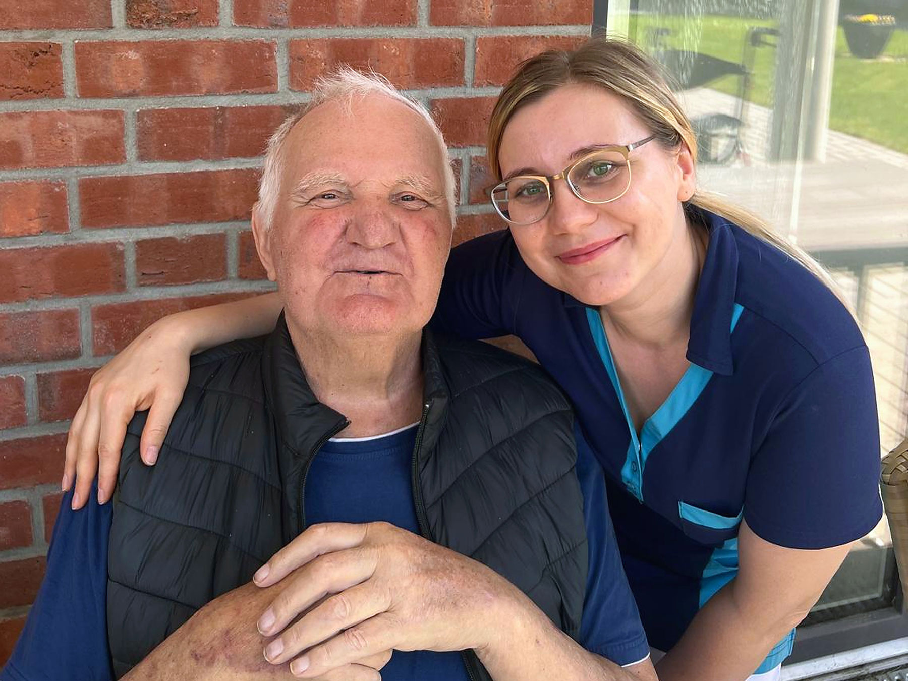 Ein älterer Mann in einer schwarzen Weste sitzt neben einer jüngeren Frau mit Brille und blauem Hemd; beide lächeln vor einer Backsteinmauer.