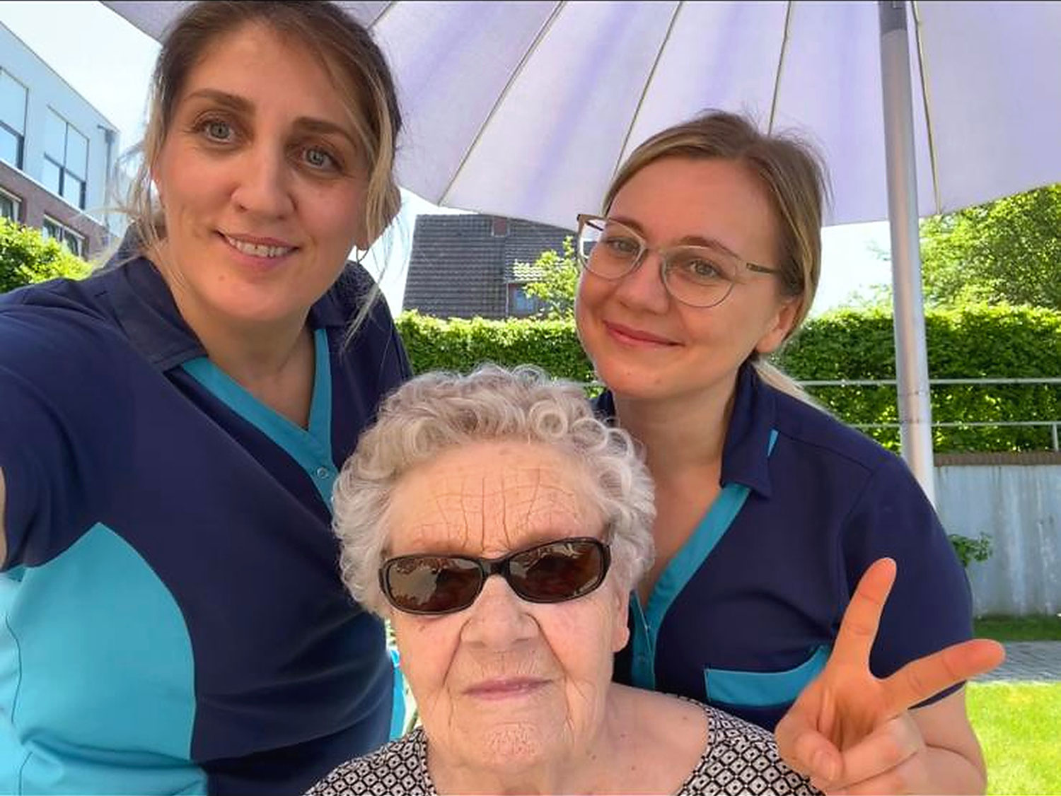 Zwei Frauen in blauen Uniformen stehen neben einer älteren Frau mit Sonnenbrille; alle lächeln im Freien unter einem Sonnenschirm. Die ältere Frau macht ein Friedenszeichen.