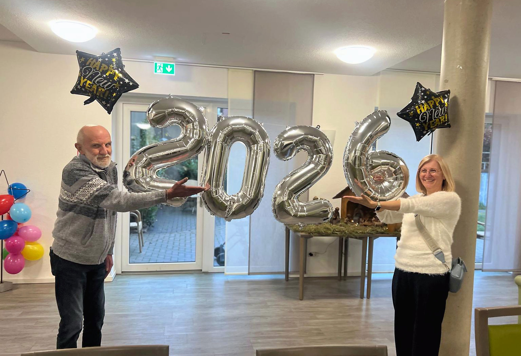 Zwei Personen halten große silberne Luftballons in den Händen, die die Zahl „2026“ bilden, im Hintergrund sind Sternluftballons mit der Aufschrift „Happy New Year“ sowie bunte Luftballondekorationen zu sehen; die Aufnahme wurde in einem Innenraum gemacht.