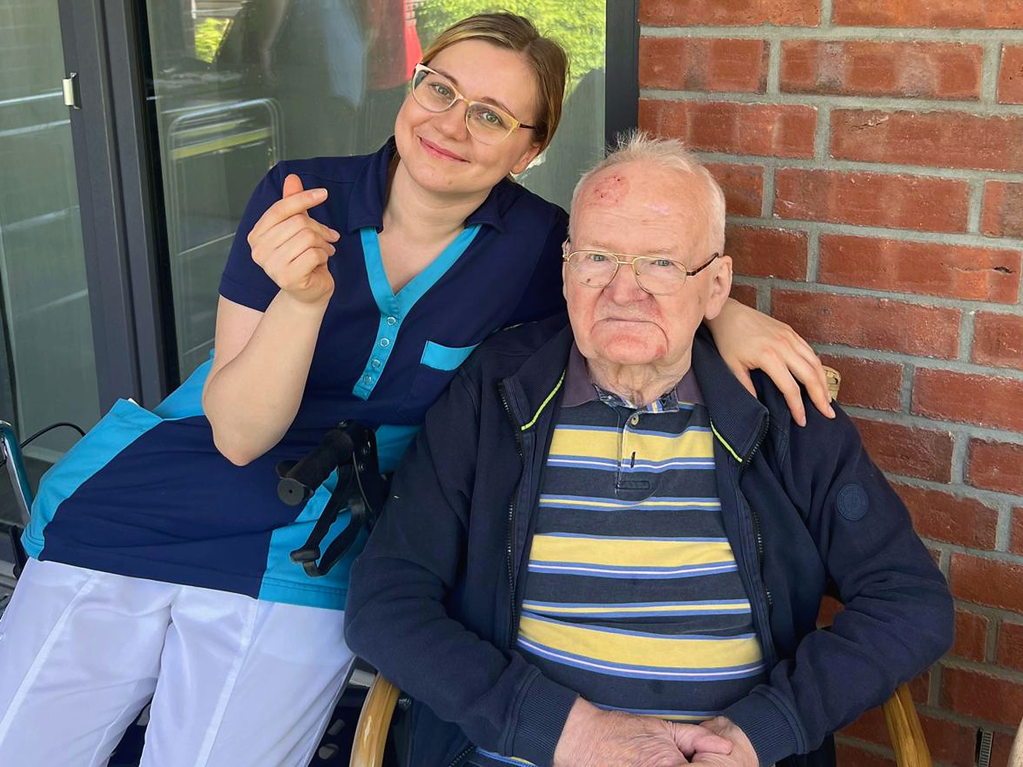 Eine Frau in medizinischer Arbeitskleidung lächelt und macht eine Handbewegung, während sie neben einem älteren Mann steht, der draußen vor einer Backsteinmauer im Rollstuhl sitzt.