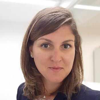 Eine Frau mit schulterlangem braunem Haar und einem marineblauen Blazer steht in einem hell erleuchteten Raum mit weißen Wänden und Glaswänden im Hintergrund.