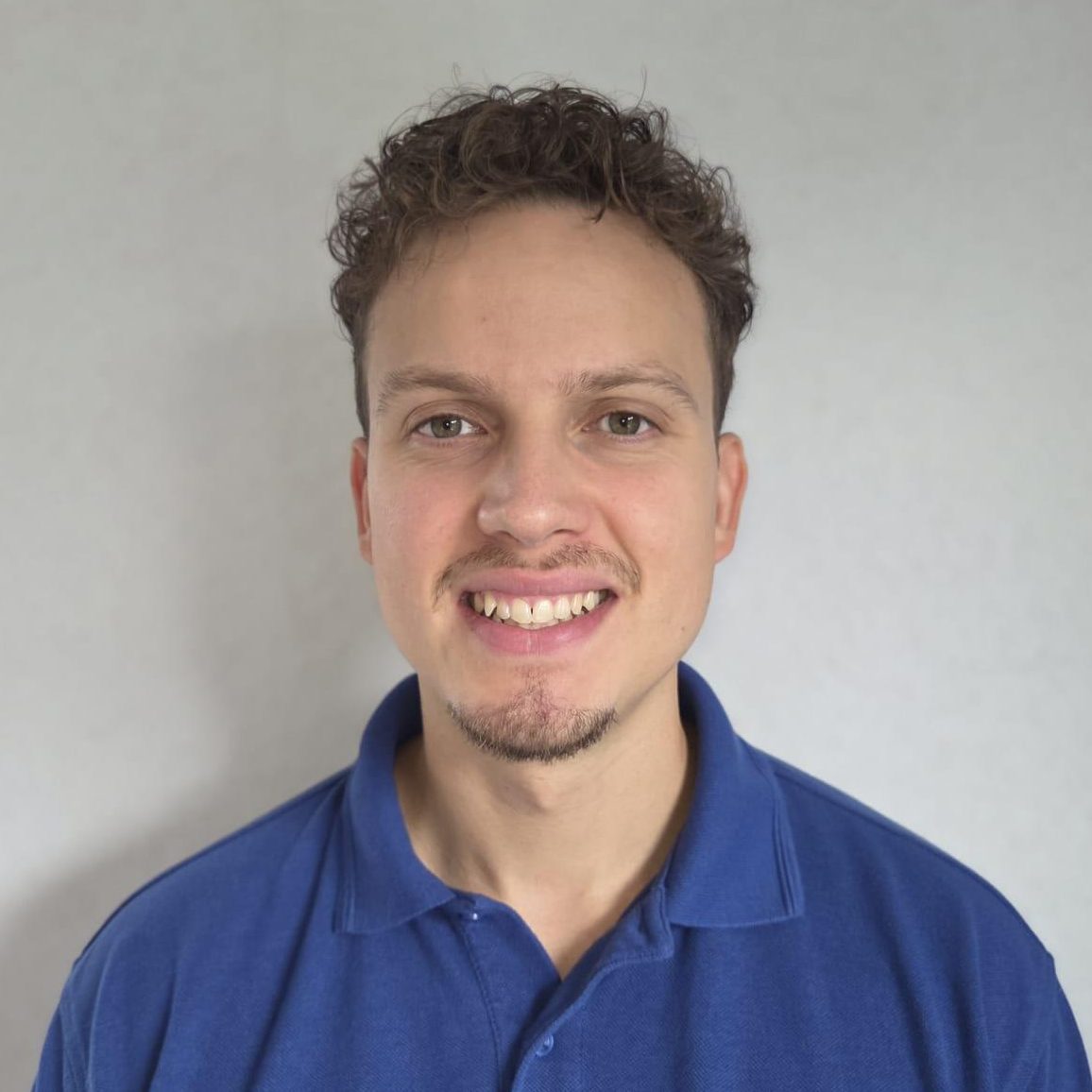 Eine Person mit kurzen Locken und einem Spitzbart lächelt in die Kamera. Sie trägt ein blaues Poloshirt mit der Aufschrift „ont“ und einem weiteren Text und steht vor einem schlichten, hellen Hintergrund.